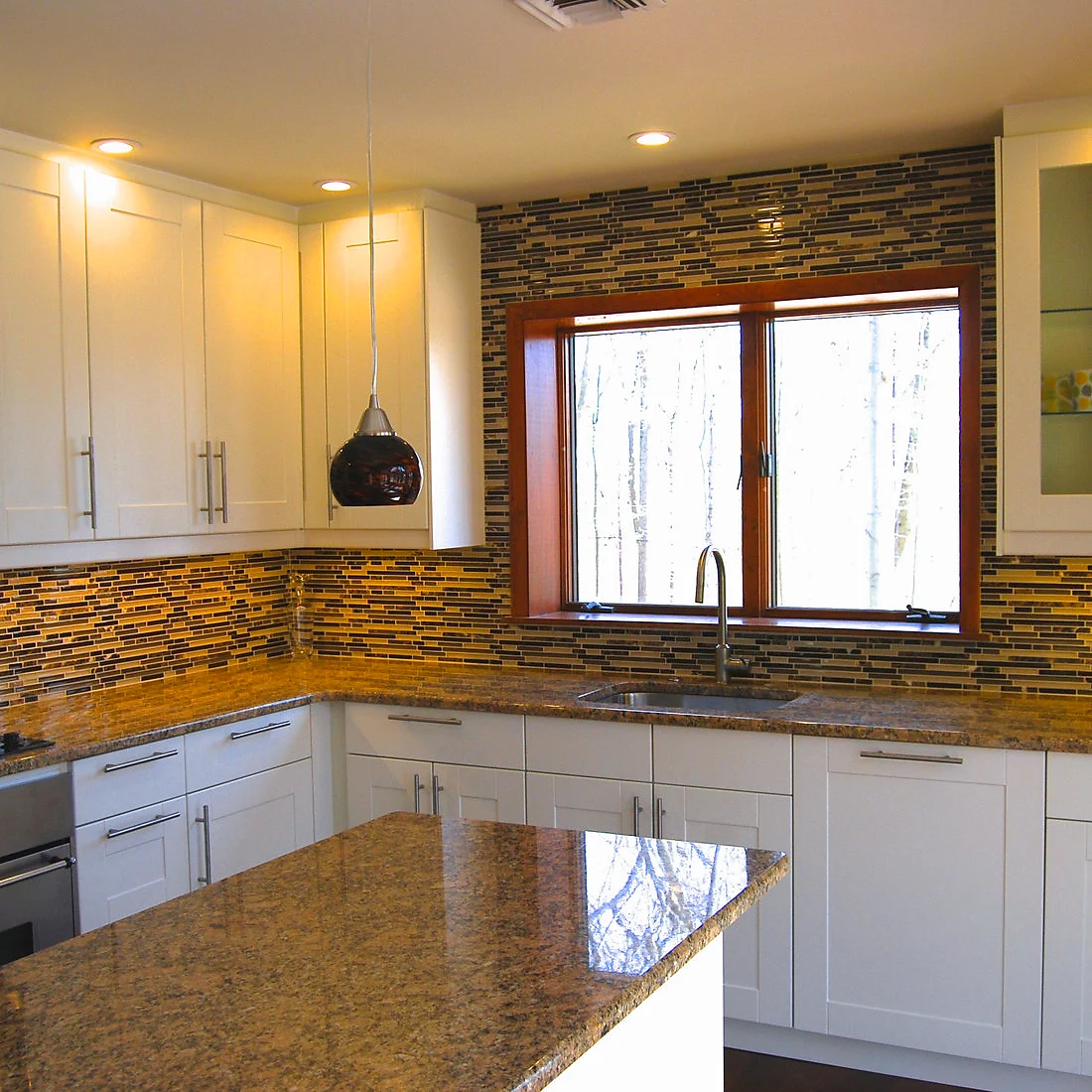 White shaker kitchen with brown glass mosaic backsplash around a wood-trimmed window, granite island in foreground