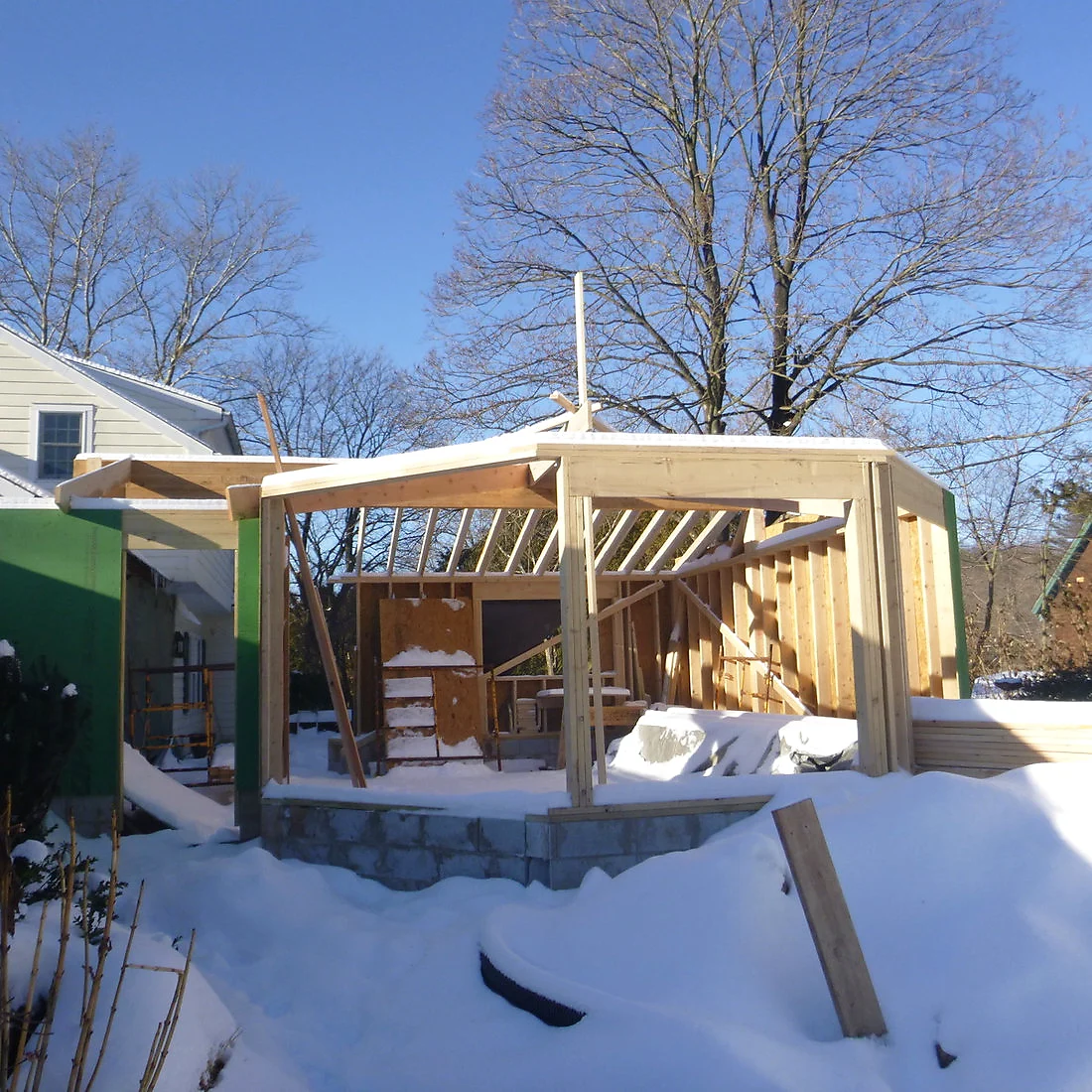 Timber-framed room addition under snow, sheathed in green weather barrier, attached to a cream clapboard house