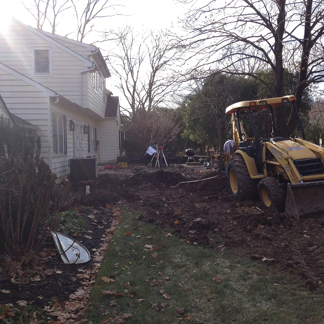 Excavation and grading alongside a cream clapboard house, backhoe cutting the footing line